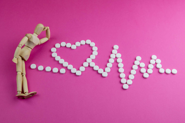Figura de juguete de madera con píldoras en forma de corazón con cardiograma sobre fondo rosa. Concepto de pastillas
. - Foto, imagen