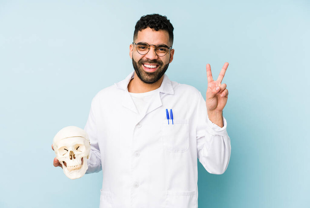 Young doctor latin man holding a skull isolated joyful and carefree showing a peace symbol with fingers. - Photo, Image