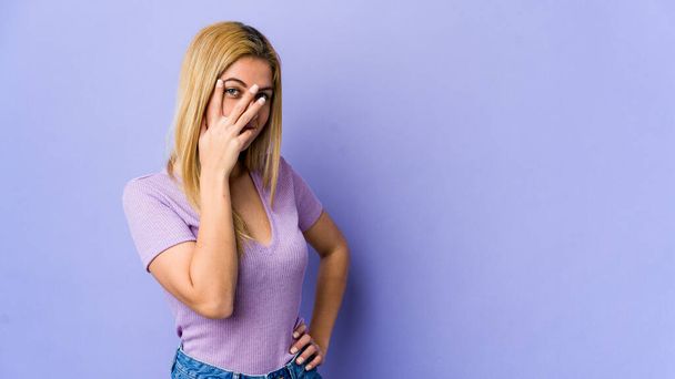 Young blonde woman isolated on purple background blink at the camera through fingers, embarrassed covering face. - Photo, Image