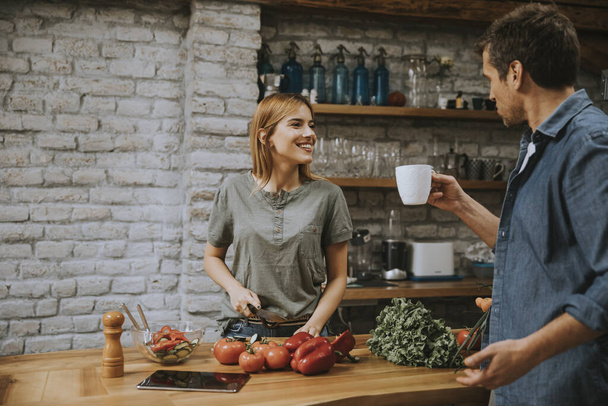 Young  woman cooking while man drinking coffee  in the rustic kitchen - Photo, Image