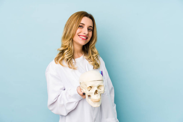 Jeune médecin femme tenant un crâne isolé regarde de côté souriant, gai et agréable
. - Photo, image