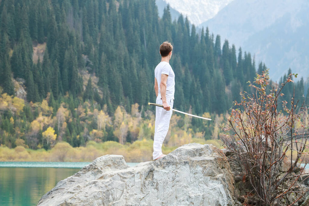 Ein junger Mann steht auf einem großen Stein und hält ein japanisches Schwert in seinen Händen. - Foto, Bild