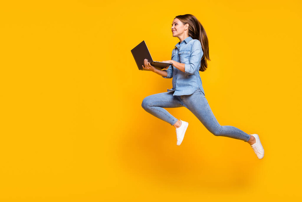 Full body profile photo of pretty business lady jump high holding notebook hands hurry work browsing laptop wear casual denim outfit white sneakers isolated yellow color background - Фото, зображення