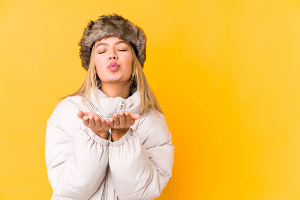Νεαρή ξανθιά καυκάσια γυναίκα που φοράει χειμωνιάτικα ρούχα απομονωμένη αναδίπλωση χειλιών και κρατώντας παλάμες για να στείλει φιλί αέρα. - Φωτογραφία, εικόνα