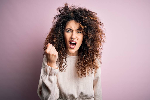 怒ったピンクの背景にカジュアルなセーターを着てピアスと巻き毛の美しい女性は怒りで叫んでいる間、拳をフラストレーションと激怒調達怒っています。激怒と積極的な概念. - 写真・画像
