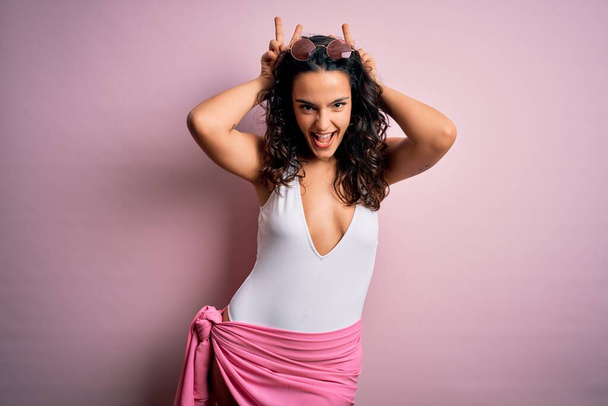 Beautiful woman with curly hair on vacation wearing white swimsuit over pink background Posing funny and crazy with fingers on head as bunny ears, smiling cheerful - Photo, Image