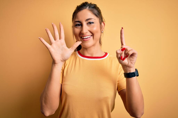 Jovem mulher atlética bonita vestindo t-shirt casual e rabo de cavalo sobre fundo amarelo mostrando e apontando para cima com os dedos número seis enquanto sorri confiante e feliz
. - Foto, Imagem