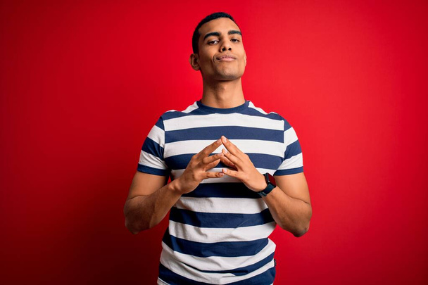 Handsome african american man wearing casual striped t-shirt standing over red background Hands together and fingers crossed smiling relaxed and cheerful. Success and optimistic - 写真・画像
