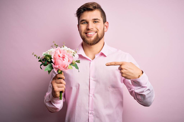 Młody blondyn romantyczny mężczyzna z brodą i niebieskimi oczami trzyma piękny bukiet kwiatów bardzo szczęśliwy wskazując ręką i palcem - Zdjęcie, obraz