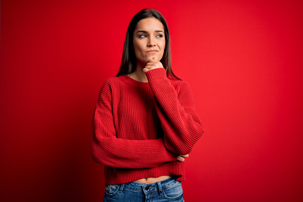 質問について考える顎の手で赤の孤立した背景に立ってカジュアルなセーターを身に着けている美しい若いブルネットの女性、集中的な表現。思慮深い顔で微笑んだ。疑いの概念. - 写真・画像