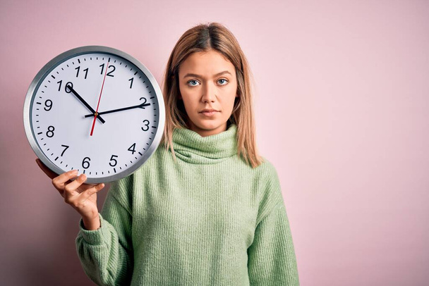 Молода красива жінка тримає годинник, що стоїть над ізольованим рожевим тлом з впевненим виразом на розумному обличчі мислення серйозно
 - Фото, зображення