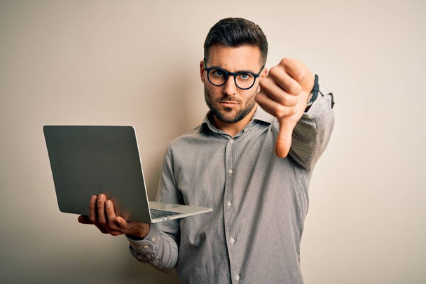 Mladý obchodník s brýlemi pracuje s počítačovým notebookem s rozzlobeným obličejem, negativní nápis ukazující odpor s palci dolů, koncept odmítnutí - Fotografie, Obrázek