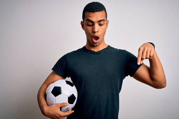 Przystojny Afroamerykanin gra w piłkę nożną trzymając piłkę na białym tle wskazując palcami pokazującymi reklamę, zaskoczoną twarz i otwarte usta - Zdjęcie, obraz