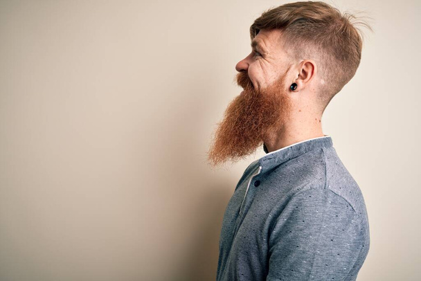 Knappe Ierse roodharige man met baard en arm tatoeage staande over geïsoleerde achtergrond op zoek naar kant, ontspannen profiel poseren met natuurlijke gezicht met zelfverzekerde glimlach. - Foto, afbeelding