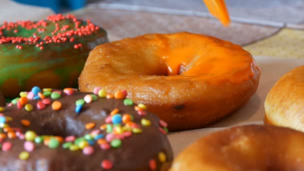 Een rij grotere ronde vers gebakken donuts op een zelfgemaakte kooktafel. Een speciale keuken siliconen borstel brengt een fel oranje glazuur aan op het oppervlak van de donut. Vet, junk food, fast food close-up - Video