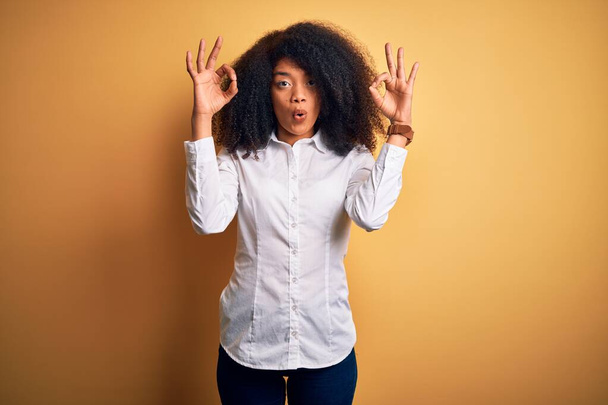 Młoda, piękna, afrykańska, elegancka kobieta z afro włosami stojącymi na żółtym tle, wyglądająca na zaskoczoną i zszokowaną, robiąc symbol aprobaty ok palcami. Szalone wyrażenie - Zdjęcie, obraz