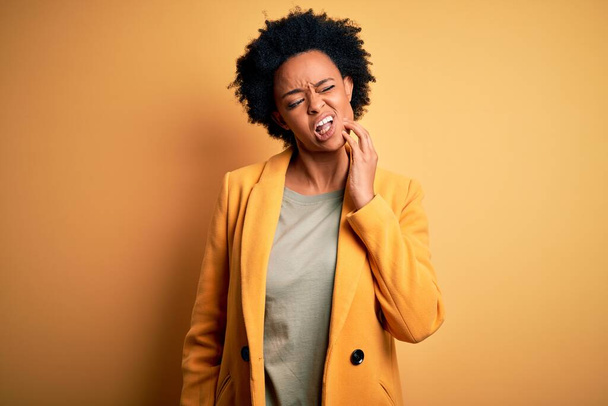 Young beautiful African American afro businesswoman with curly hair wearing yellow jacket touching mouth with hand with painful expression because of toothache or dental illness on teeth. Dentist - Foto, Imagen