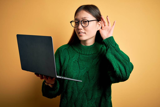 Junge asiatische Geschäftsfrau mit Brille und Computer-Laptop, die lächelnd mit der Hand über dem Ohr Gerüchten oder Klatsch lauscht. Konzept der Gehörlosigkeit. - Foto, Bild