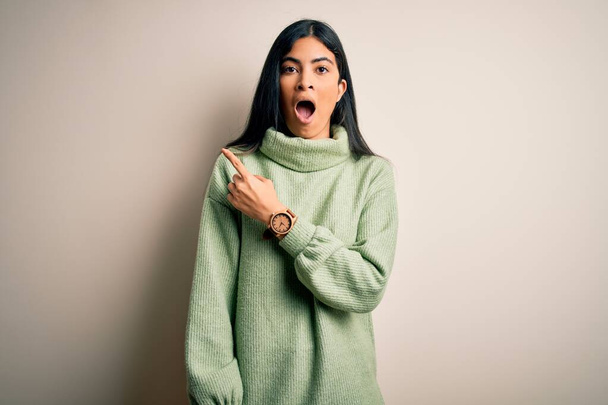 Young beautiful hispanic woman wearing green winter sweater over isolated background Surprised pointing with finger to the side, open mouth amazed expression. - Фото, зображення