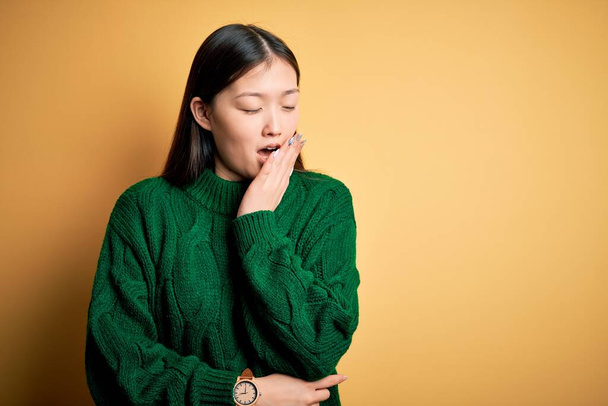 Nuori kaunis aasialainen nainen yllään vihreä talvi villapaita yli keltainen eristetty tausta kyllästynyt haukottelu väsynyt peittää suun käsin. Levottomuus ja uneliaisuus
. - Valokuva, kuva