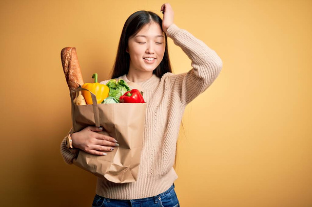 Joven mujer asiática sosteniendo bolsa de papel de comestibles sanos frescos sobre fondo aislado amarillo sonriendo confiado tocando el cabello con la mano hacia arriba gesto, posando atractivo y de moda
 - Foto, Imagen