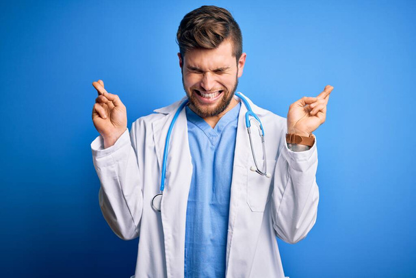 Jonge blonde dokter man met baard en blauwe ogen met witte jas en stethoscoop gekruiste vinger glimlachend met hoop en ogen dicht. Geluk en bijgelovig concept. - Foto, afbeelding