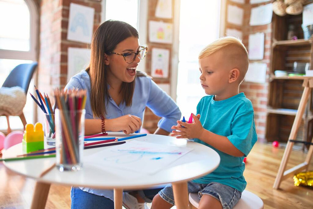 Genç beyaz çocuk öğretmeniyle oyun okulunda oynuyor. Anne ve oğlu oyun odasında renkli kalemlerle beraberlik çiziyor. - Fotoğraf, Görsel