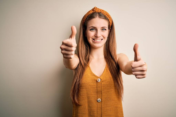 Молодая красивая рыжая женщина в повседневной футболке и диадеме на жёлтом фоне, одобряющая позитивный жест рукой, улыбается и радуется успеху. Жест победителя
. - Фото, изображение