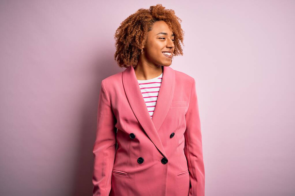 Молодая красивая афро-американская деловая женщина с вьющимися волосами в элегантной розовой куртке смотрит в сторону с улыбкой на лице, естественное выражение лица. Смеясь уверенно
. - Фото, изображение