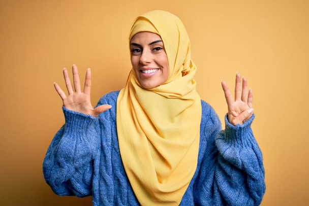 Genç, güzel, esmer, Müslüman kadın izole edilmiş sarı arka plan üzerine Arap başörtüsü takıyor ve dokuz numarayı işaret ederek gülümsüyor.. - Fotoğraf, Görsel