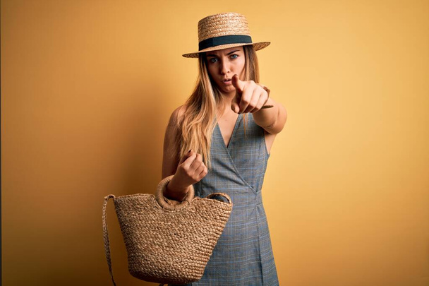 Mulher loira bonita com olhos azuis em férias usando vestido e chapéu de verão segurando saco apontando com o dedo para a câmera e para você, sinal de mão, gesto positivo e confiante da frente
 - Foto, Imagem