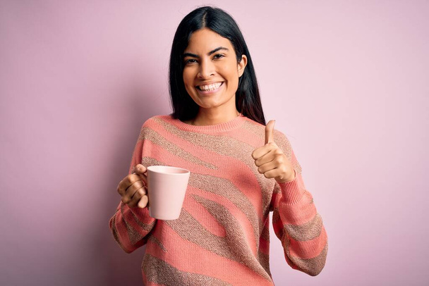 若い美しいですヒスパニック系女性飲む上のホットコーヒーカップのピンク隔離された背景幸せなで大きな笑顔を行いますokサイン,指で親指アップ,優れたサイン - 写真・画像