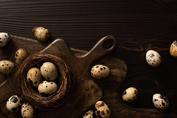 top view of quail eggs in nest and on brown cutting boards on dark wooden surface - Foto, Imagen