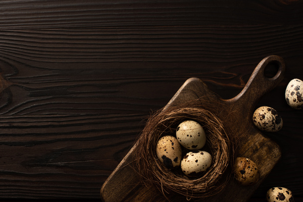 top view of quail eggs in nest on brown cutting board on dark wooden background - Zdjęcie, obraz