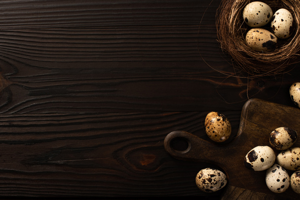 top view of quail eggs in brown nest and on chopping board on dark wooden background - Zdjęcie, obraz