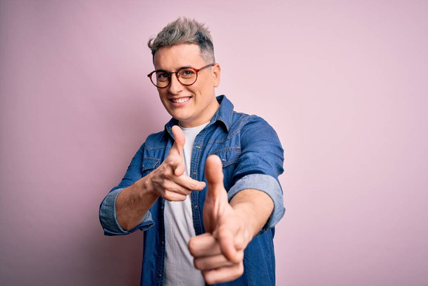Jonge knappe moderne man draagt bril en spijkerjasje over roze geïsoleerde achtergrond wijzende vingers naar camera met vrolijk en grappig gezicht. Goede energie en vibraties. - Foto, afbeelding