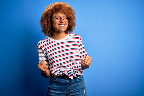 Afrikkalainen amerikkalainen nainen, jolla on kiharat hiukset lomalla yllään kesähattu ja raidallinen t-paita erittäin onnellinen ja innoissaan tekemässä voittajan elettä kädet kohotettuina, hymyillen ja huutaen menestystä. Juhlan käsite
. - Valokuva, kuva