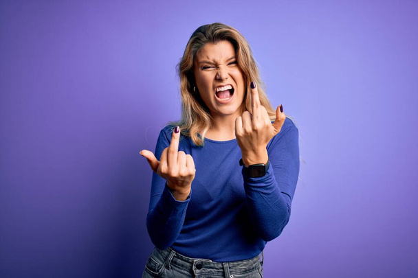 Young beautiful blonde woman wearing casual t-shirt over isolated purple background Showing middle finger doing fuck you bad expression, provocation and rude attitude. Screaming excited - Foto, Bild