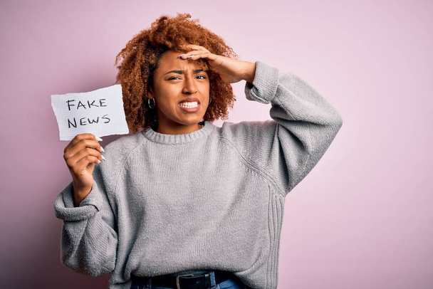 Fiatal afro-amerikai afro nő göndör haj tartó papír hamis hírek üzenetet hangsúlyozta kezét a fejre, sokkolt a szégyen és meglepetés arc, dühös és frusztrált. Félelem és tévedés. - Fotó, kép