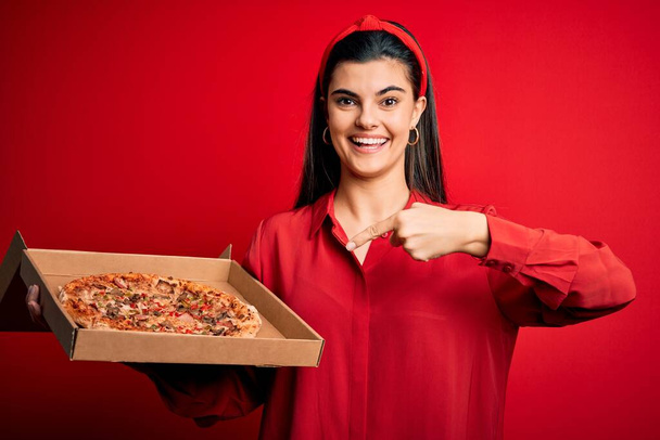 Genç, güzel esmer bir kadın, elinde İtalyan pizzası olan bir teslimat kutusu tutuyor. Kırmızı arka planda, mutlu bir şekilde eli ve parmağı işaret ediyor. - Fotoğraf, Görsel