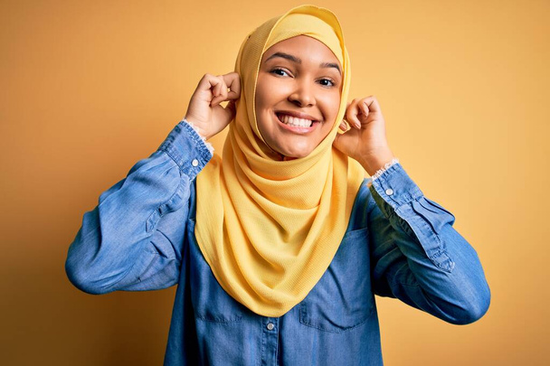 Молода красива жінка з кучерявим волоссям носить арабський традиційний хіджаб на жовтому тлі Посміхаючись тягнути вуха пальцями, смішний жест. Проблема прослуховування
 - Фото, зображення
