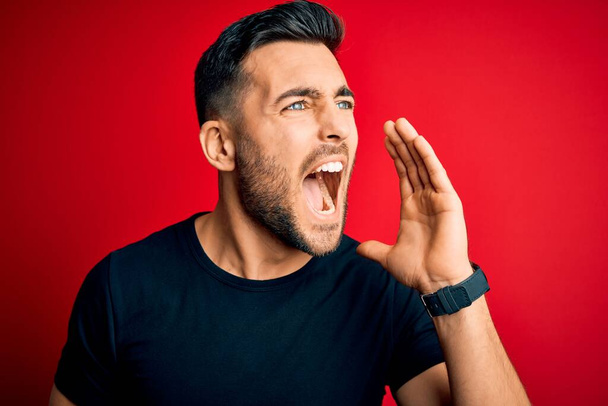 Young handsome man wearing casual black t-shirt standing over isolated red background shouting and screaming loud to side with hand on mouth. Communication concept. - Photo, image