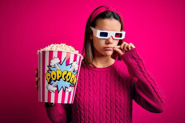 Young beautiful girl watching movie using 3d glasses eating box with popcorns mouth and lips shut as zip with fingers. Secret and silent, taboo talking - Photo, Image
