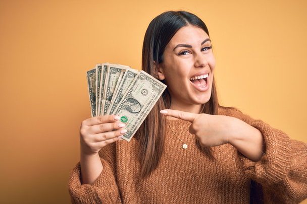 Jovem mulher bonita segurando dólares em pé sobre fundo laranja isolado muito feliz apontando com a mão e o dedo
 - Foto, Imagem