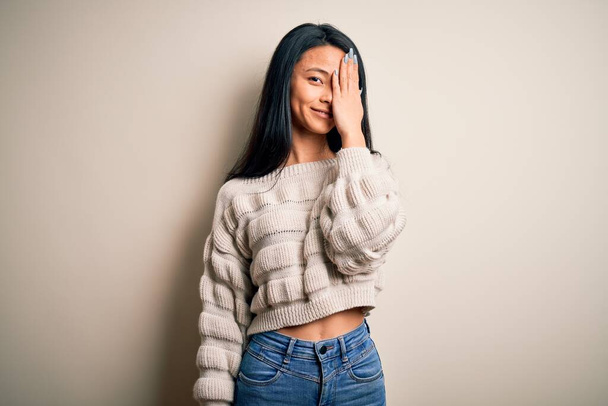 Jonge mooie Chinese vrouw dragen casual trui over geïsoleerde witte achtergrond bedekken een oog met de hand, zelfverzekerde glimlach op het gezicht en verrassing emotie. - Foto, afbeelding