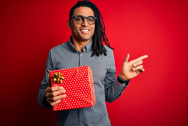 Афроамериканец с дредами держит настоящую коробку на красном изолированном фоне очень доволен указывая рукой и пальцем в сторону
 - Фото, изображение