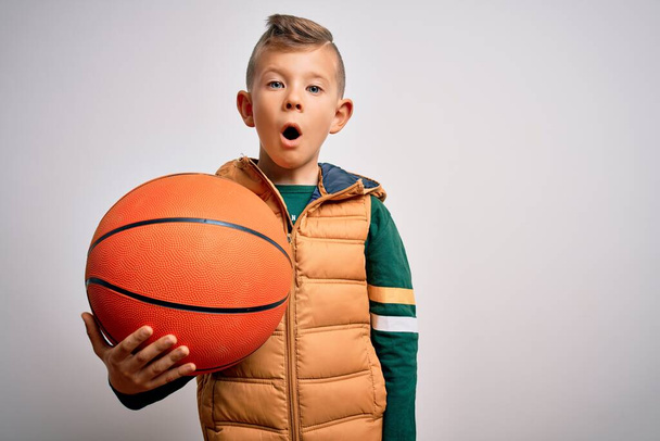 Молодий маленький білий спортивний хлопчик грає в баскетбол, тримаючи помаранчевий м'яч над ізольованим тлом, наляканий шоком з несподіваним обличчям, боїться і схвильований виразом страху
 - Фото, зображення