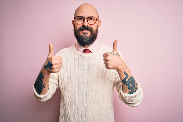 Рукоплечий лысый мужчина с бородой и татуировкой в очках и свитере на розовом фоне делает позитивный жест рукой, улыбается и радуется. Веселая экспрессия и жест победителя. - Фото, изображение