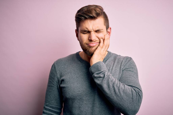 Young handsome blond man with beard and blue eyes wearing casual sweater touching mouth with hand with painful expression because of toothache or dental illness on teeth. Dentist - Foto, Imagem