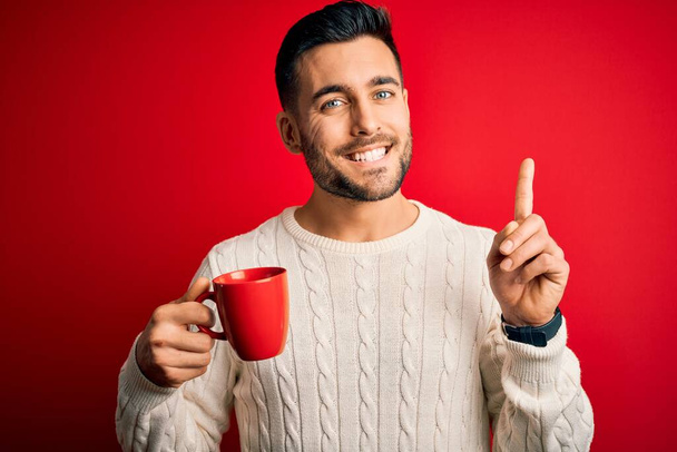 Genç yakışıklı bir adam kırmızı izole arka planda bir fincan sıcak kahve içiyor. Bir fikir ya da soru işareti ile şaşırmış, mutlu bir yüz, bir numara. - Fotoğraf, Görsel